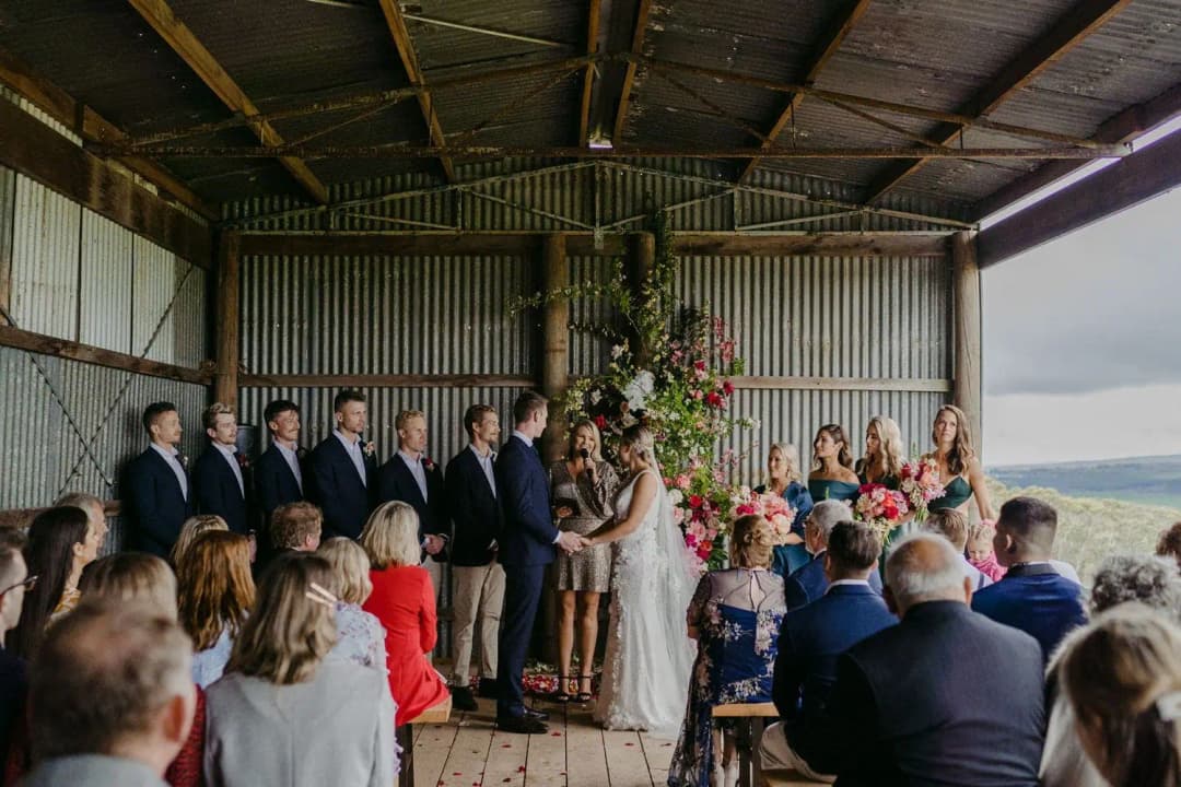 Couple and wedding party standing before seated guests inside a rustic shed.