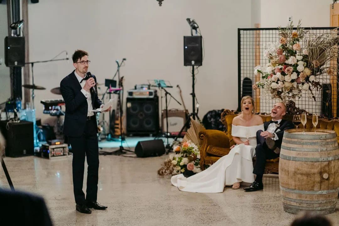 Man giving a microphone speech before the seated couple in a reception room.