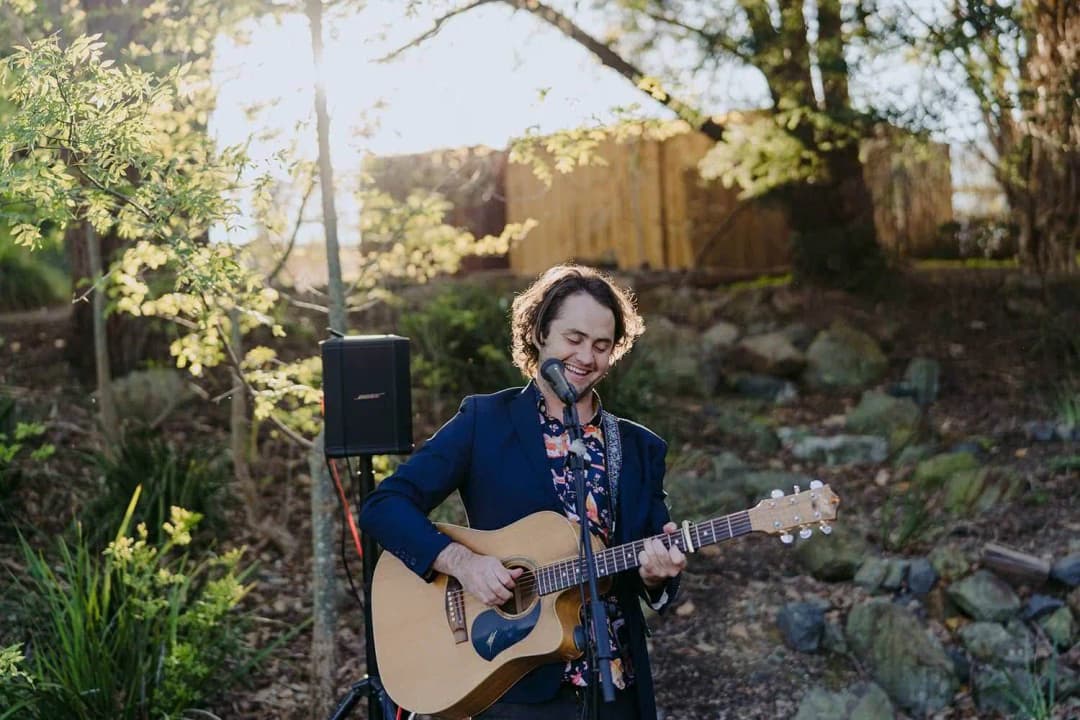 Guitarist singing into a microphone beside garden rocks and trees.