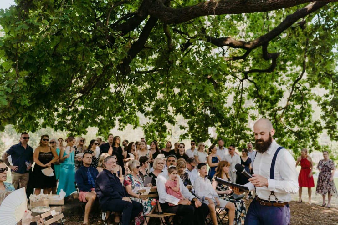 Celebrant standing with notes beside guests seated beneath a leafy tree