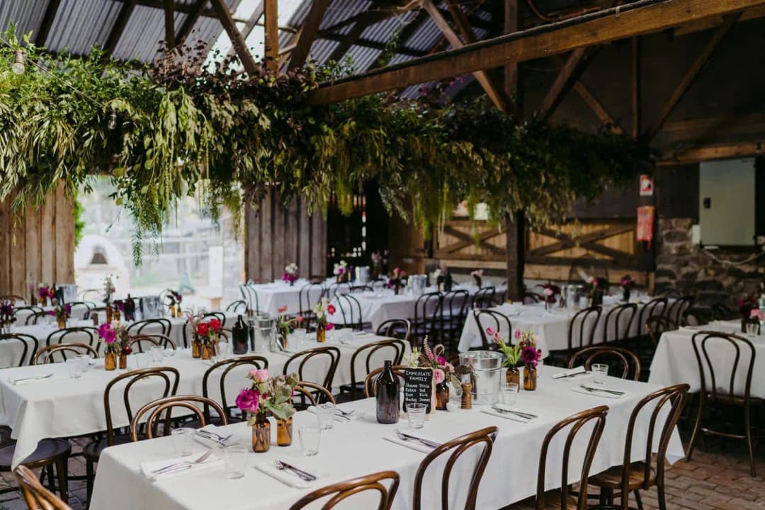 Barn reception tables arranged beneath exposed rafters and hanging foliage