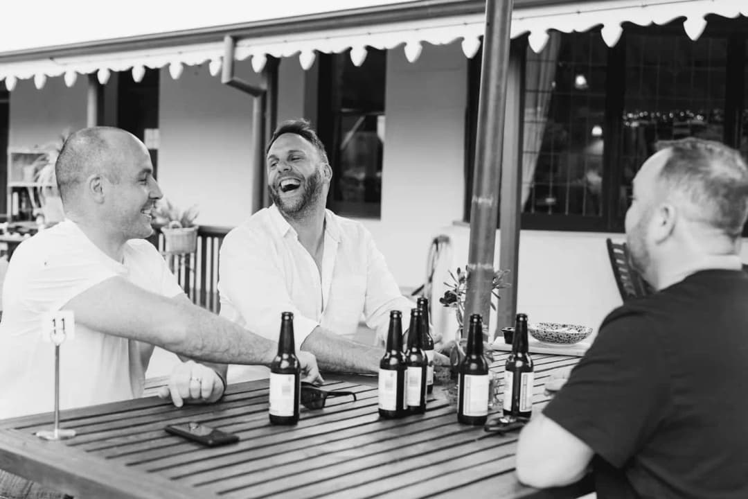 Black and white photograph of men laughing around a table with beer bottles