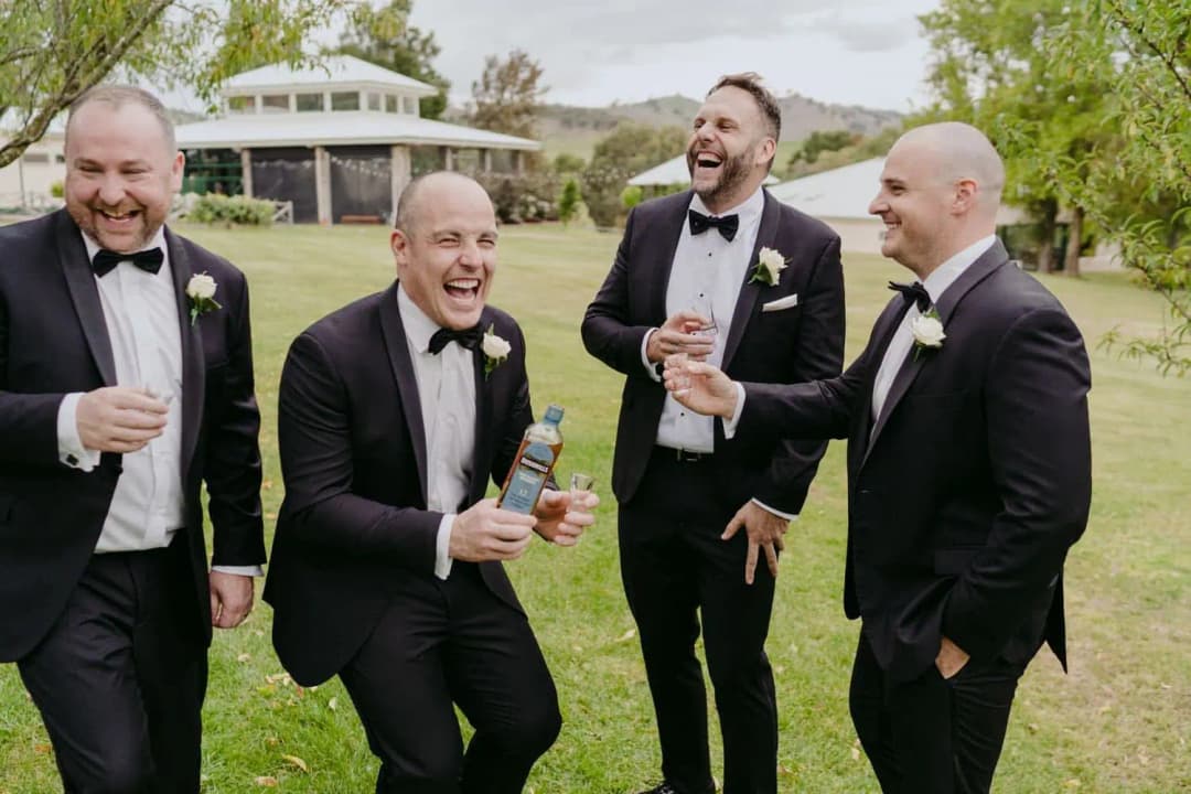 Four men in tuxedos laughing together on a lawn with drinks