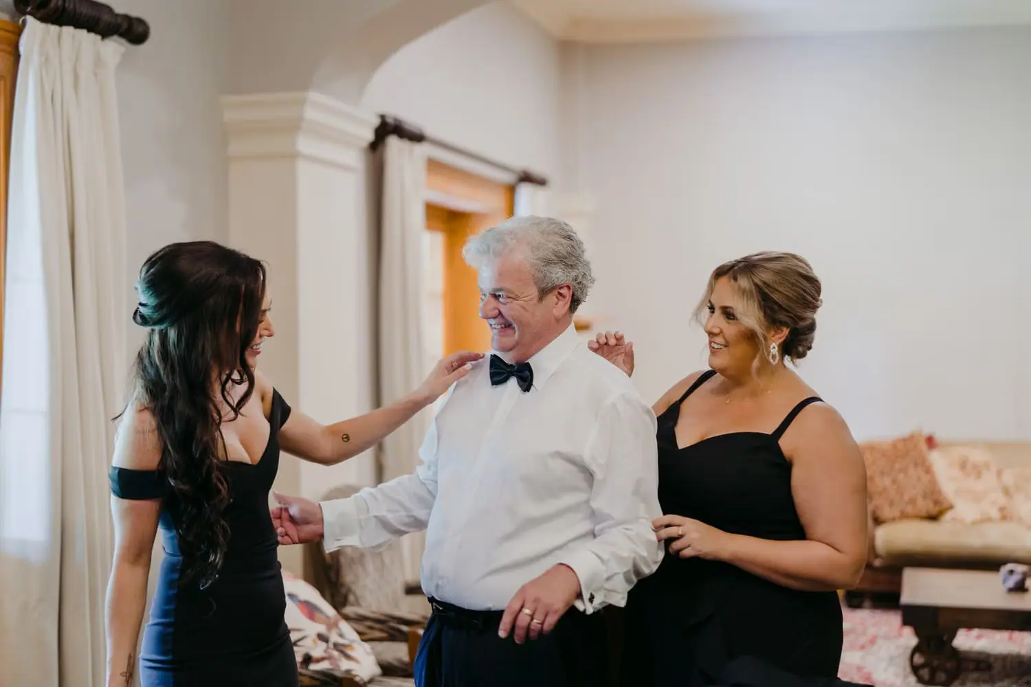 Two women in black dresses greeting a grey-haired man wearing a bow tie