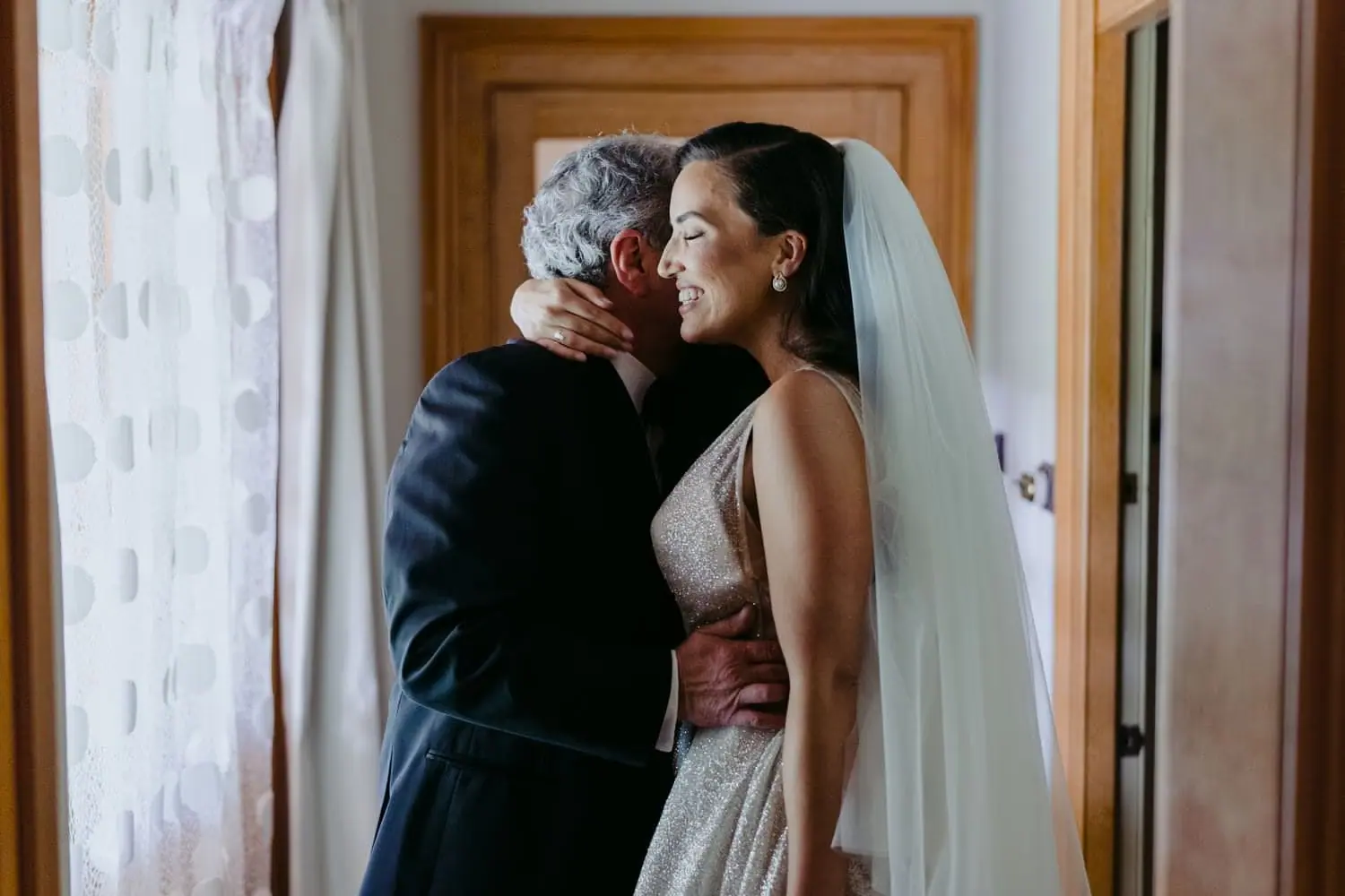Bride embracing a grey-haired man beside a curtained window