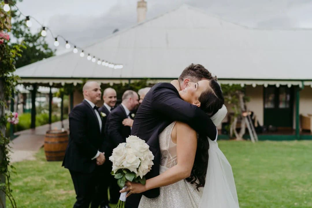 Bride and groom kissing while groomsmen watch beneath outdoor fairy lights