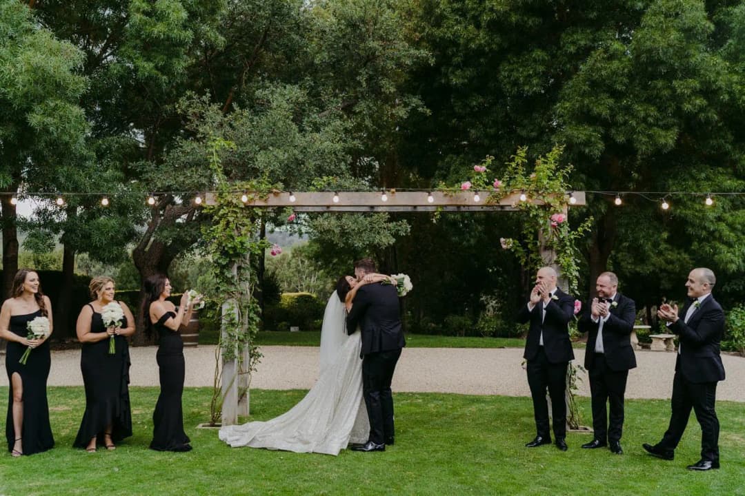 Wedding party applauding as the bride and groom embrace beneath a timber floral arch