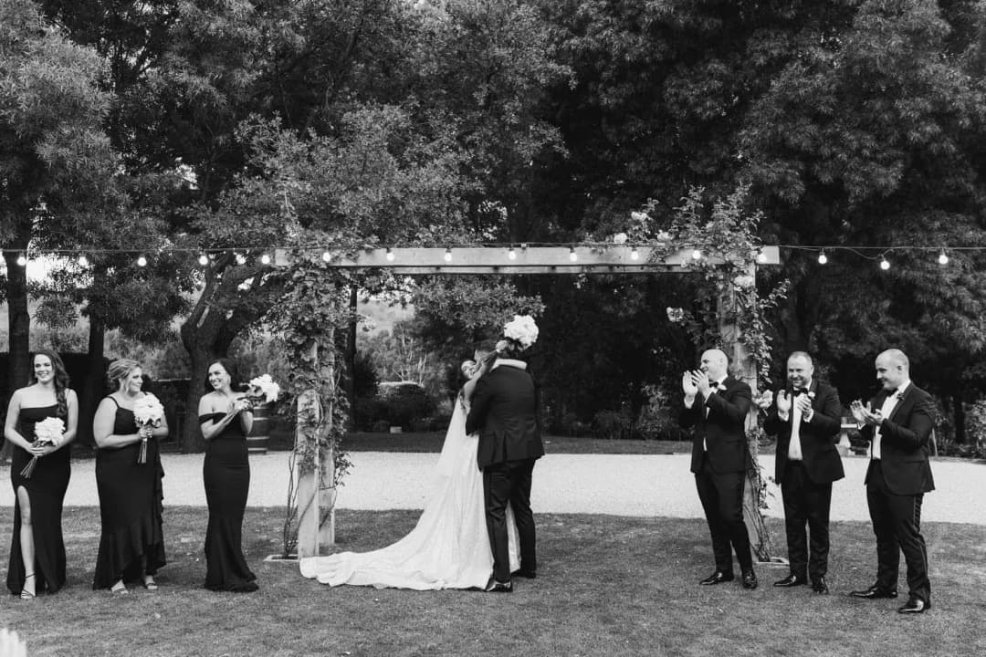 Black and white photograph of the wedding party applauding the bride and groom beneath an arch