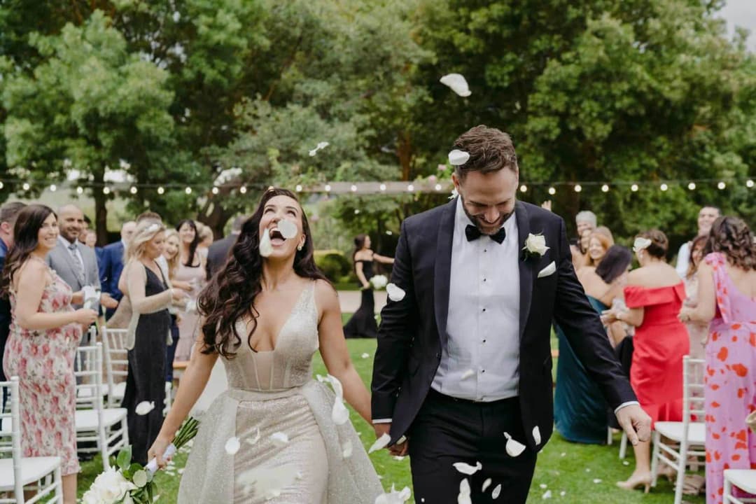 Bride and groom smiling as petals fall while they walk past cheering guests