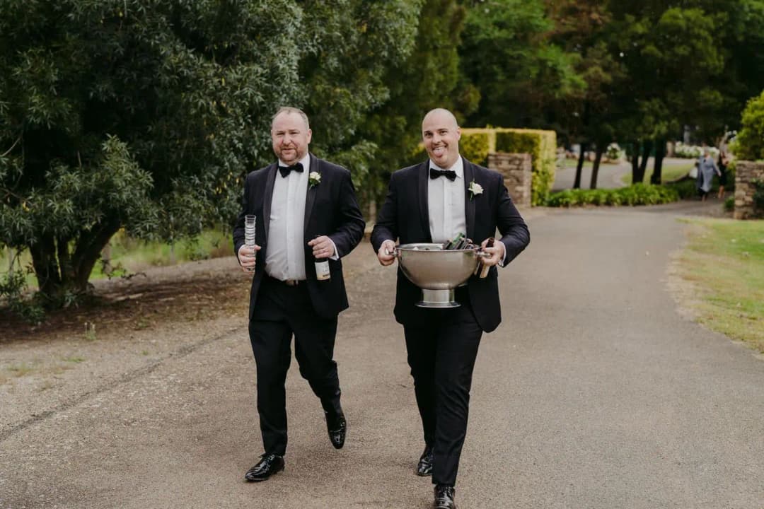 Two men in tuxedos walking along a driveway with drinks and a metal ice bucket