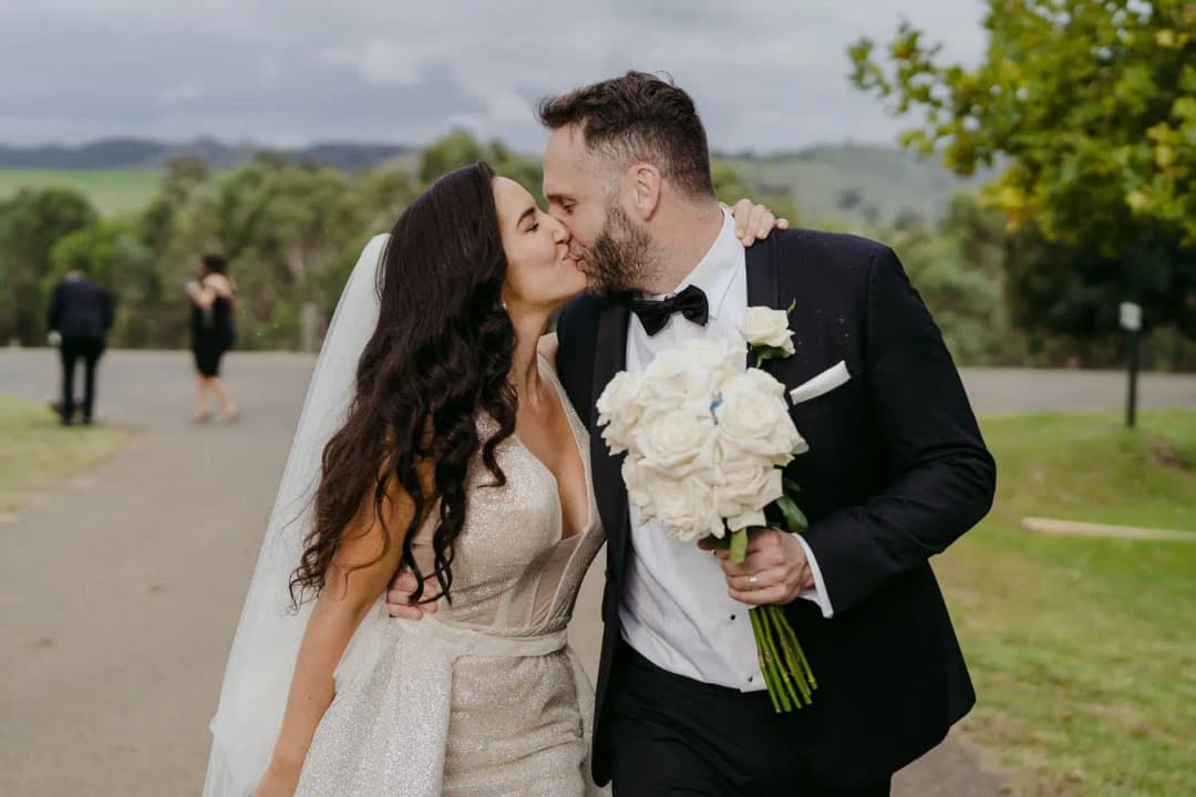 Bride and groom kissing beside a tree-lined driveway while holding a white bouquet