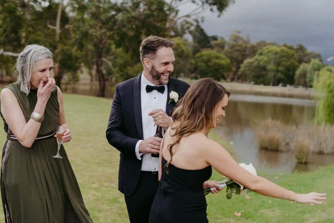 Groom and two guests laughing beside a lake with drinks