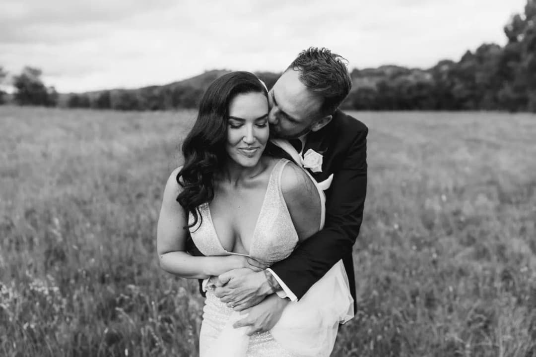 Black and white photograph of the groom embracing the bride from behind in a grassy field