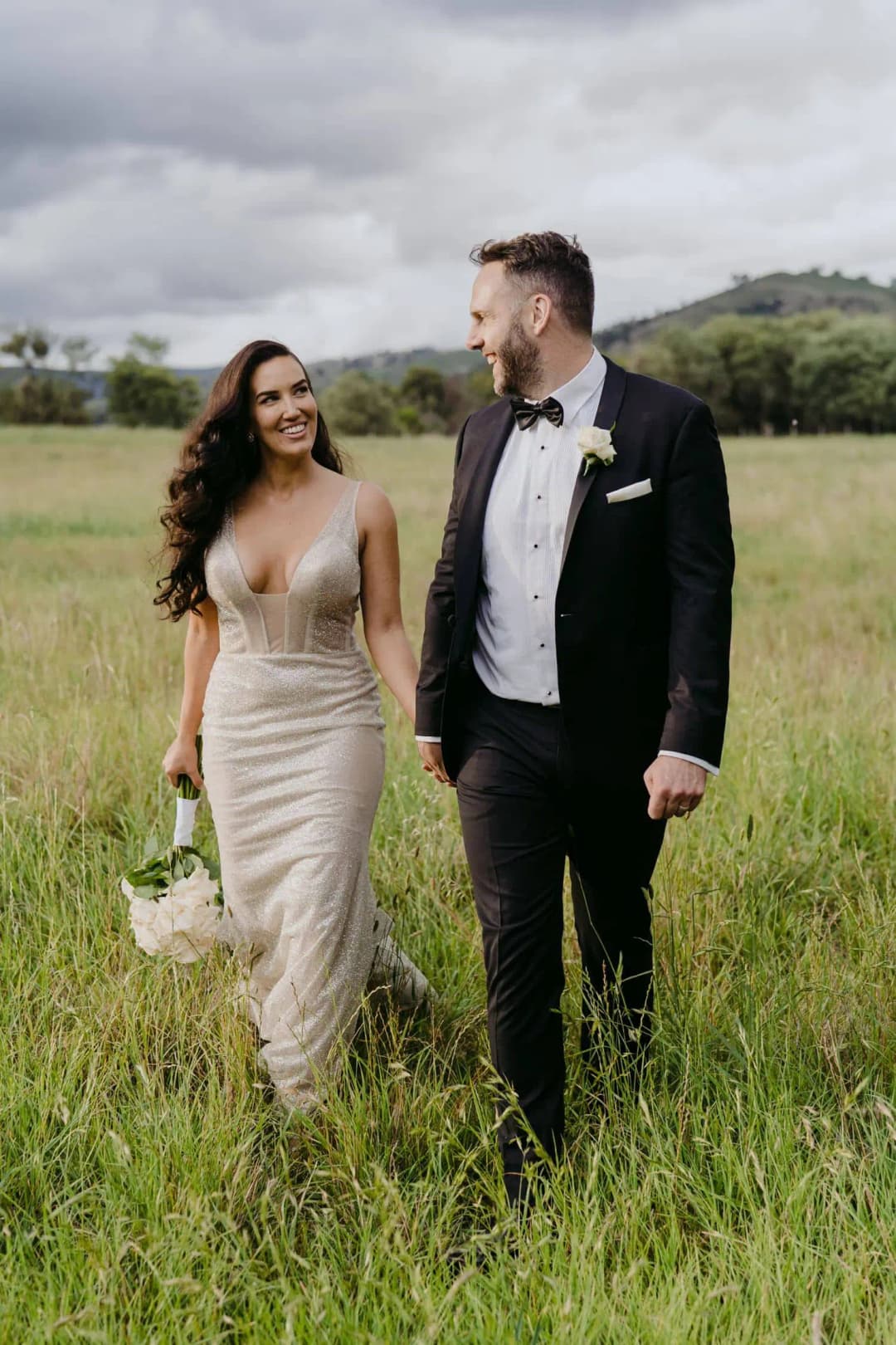 Bride and groom walking together through a grassy field beneath a cloudy sky