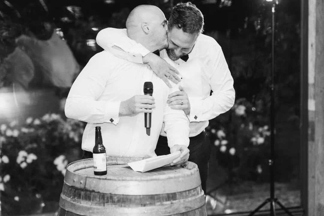 Black and white photograph of a man hugging the groom while holding a microphone beside a barrel