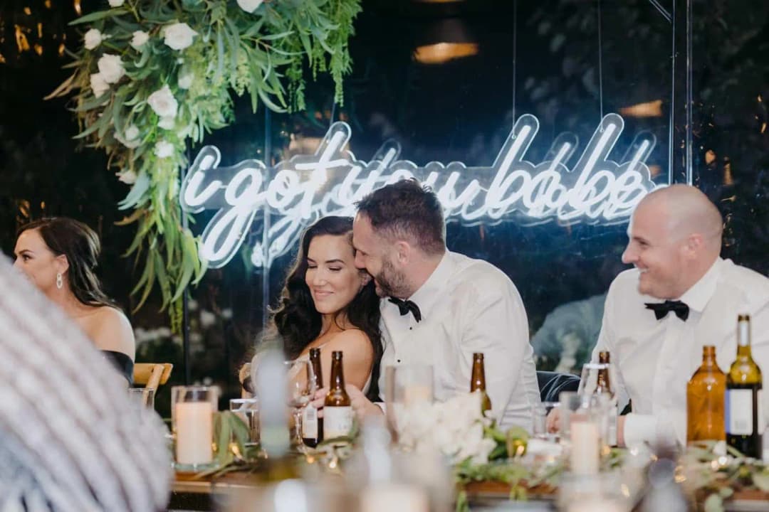 Groom leaning toward the bride at the reception table beneath an illuminated sign