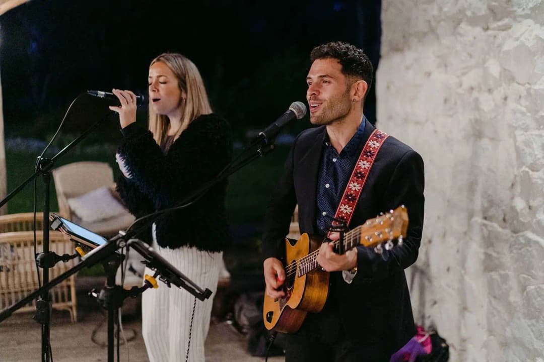 Singer and guitarist performing beside a pale stone wall.