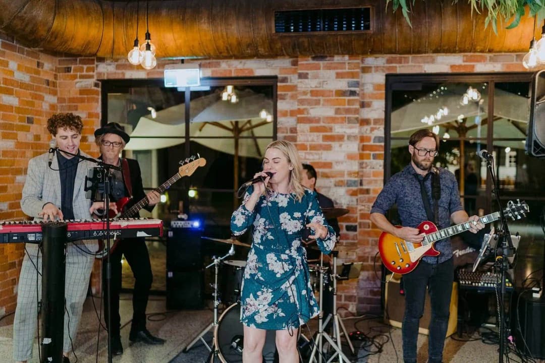 Band performing with a singer, keyboard and guitar beside a brick wall.
