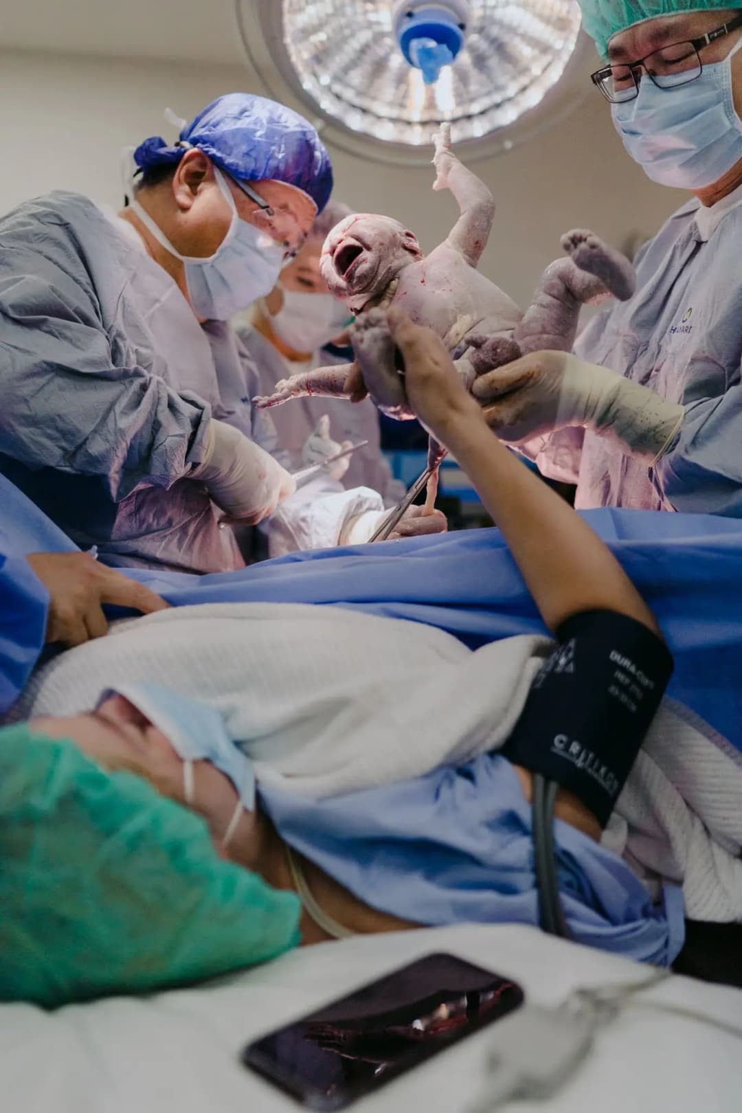 Medical staff holds a newborn above a patient reaching up from a hospital bed.