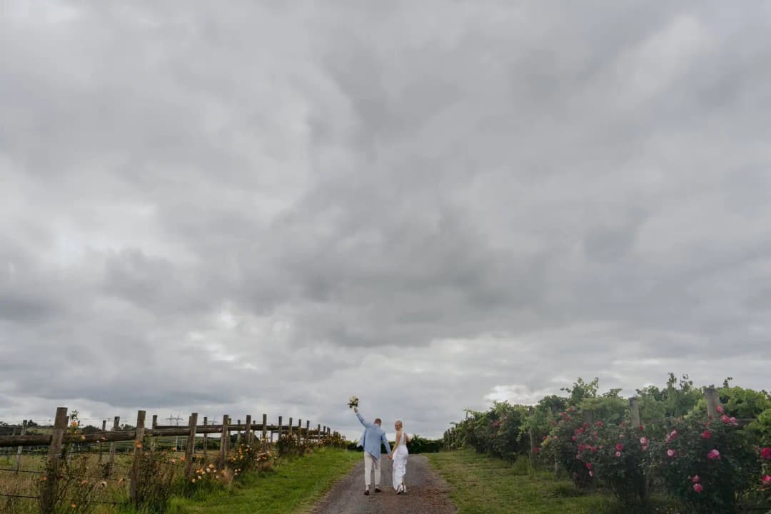 Distant couple walks along a vineyard path beneath a broad cloudy sky.