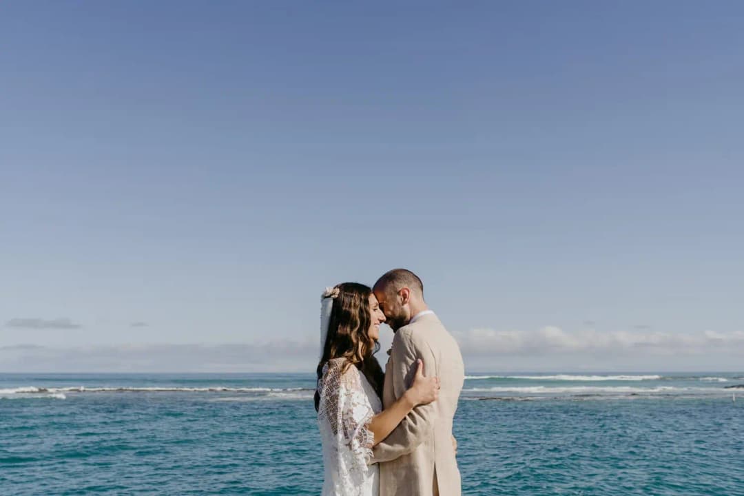 Couple embraces beside the blue ocean under a clear sky.