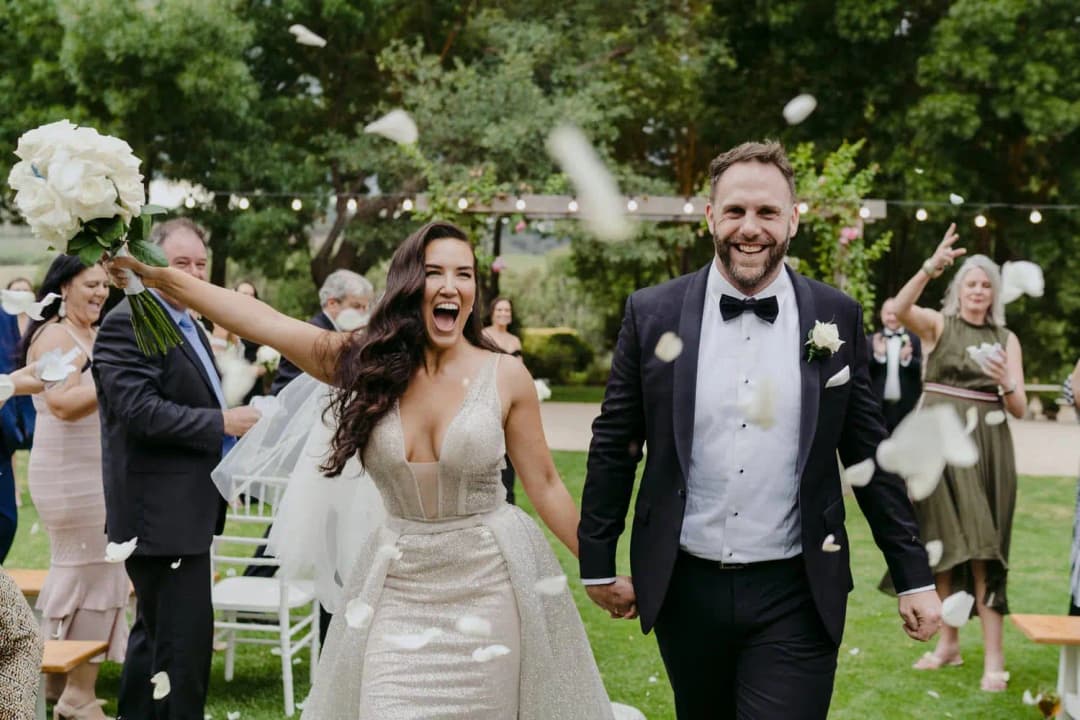 Newlyweds smile as guests shower them with confetti on a garden lawn.