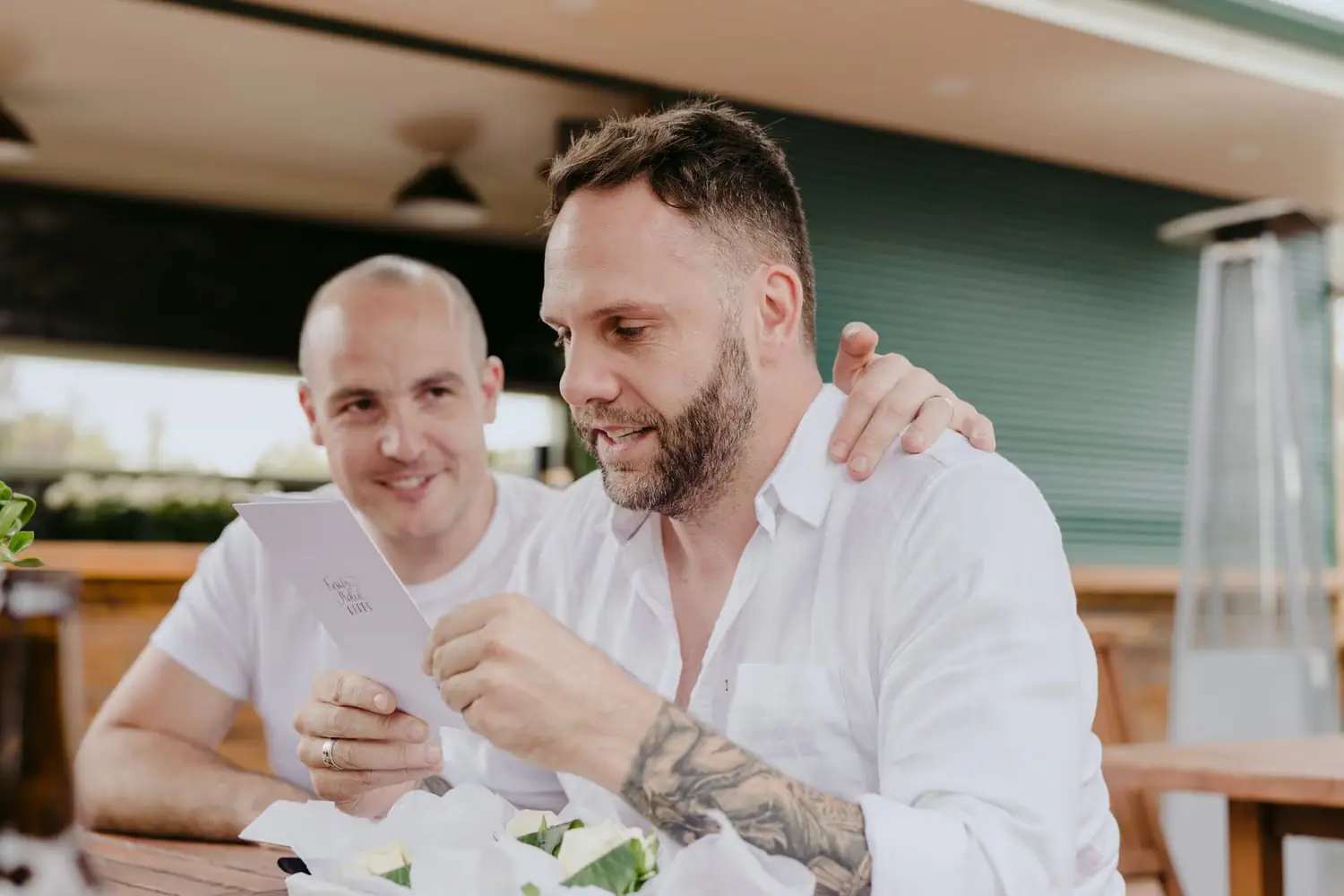Two men smile together at a reception table with their arms around each other.