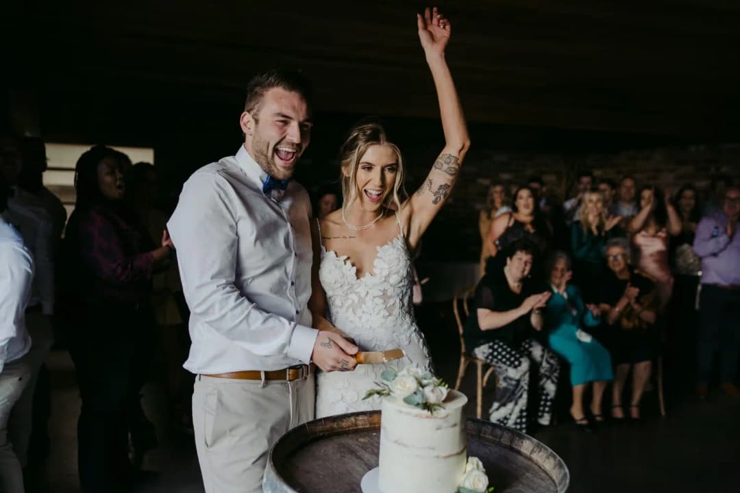 Bride raises an arm while the couple cuts their wedding cake before guests.