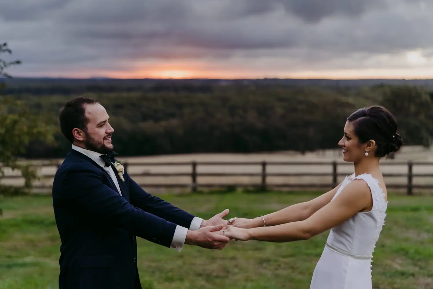 Couple holds both hands facing each other on a lawn at sunset.