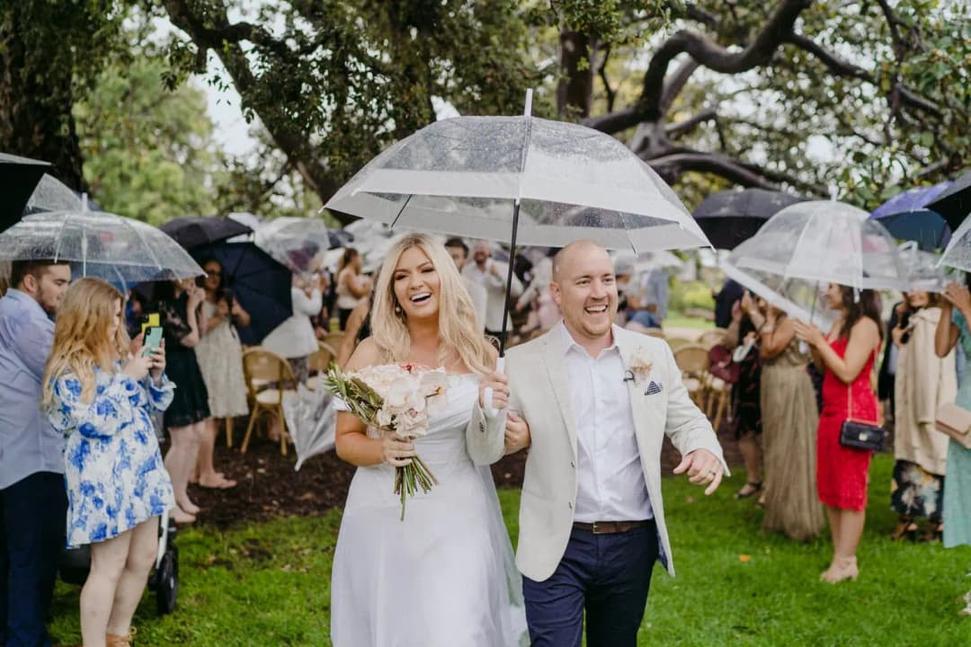 Couple walks through guests holding clear umbrellas in a rainy garden.