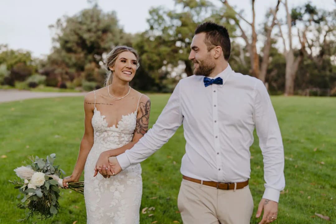 Couple walks hand in hand across a lawn holding a white bouquet.
