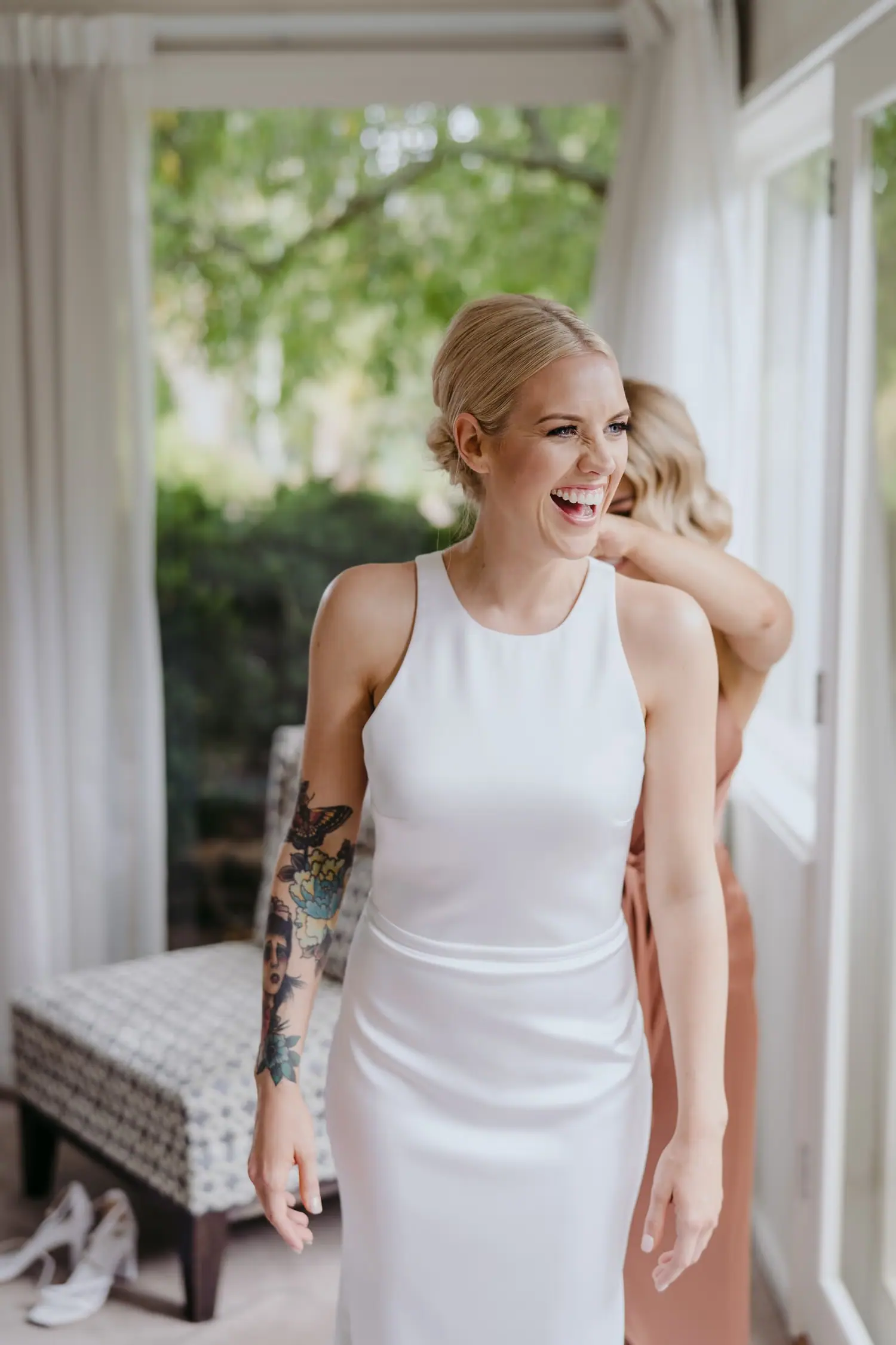 Bride laughs while a friend helps with her dress beside a window.
