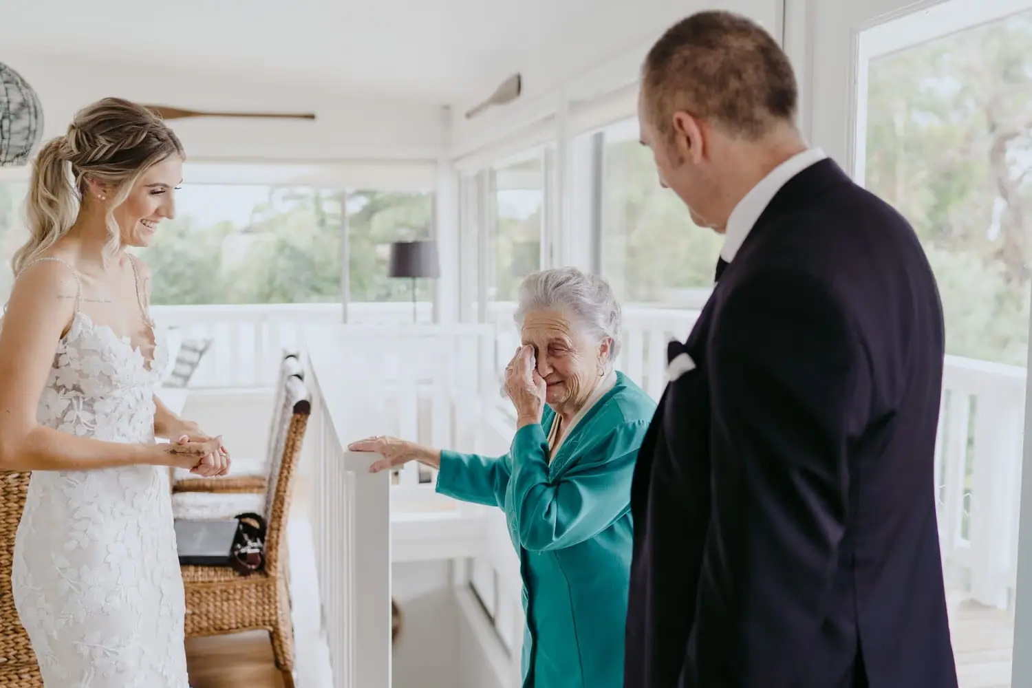 Bride and groom smile toward an older woman in a bright room.