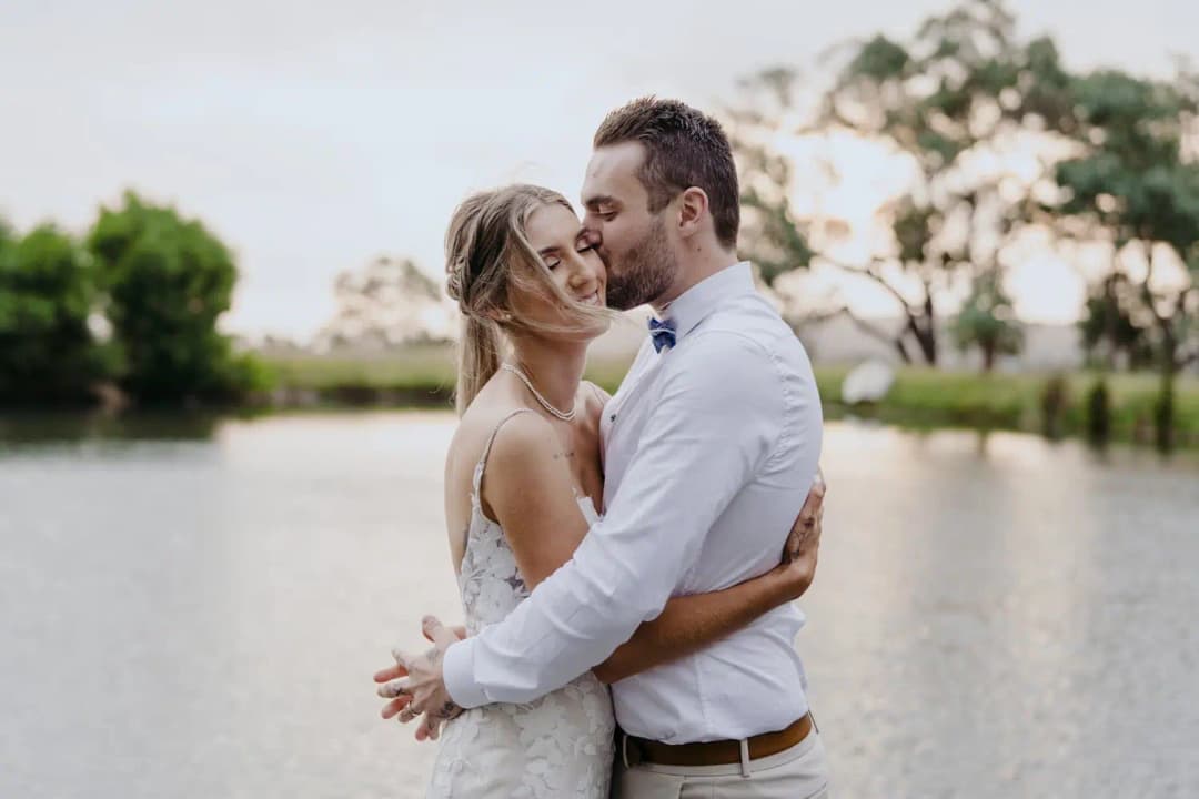 Couple kisses beside a pond with trees beyond the water.