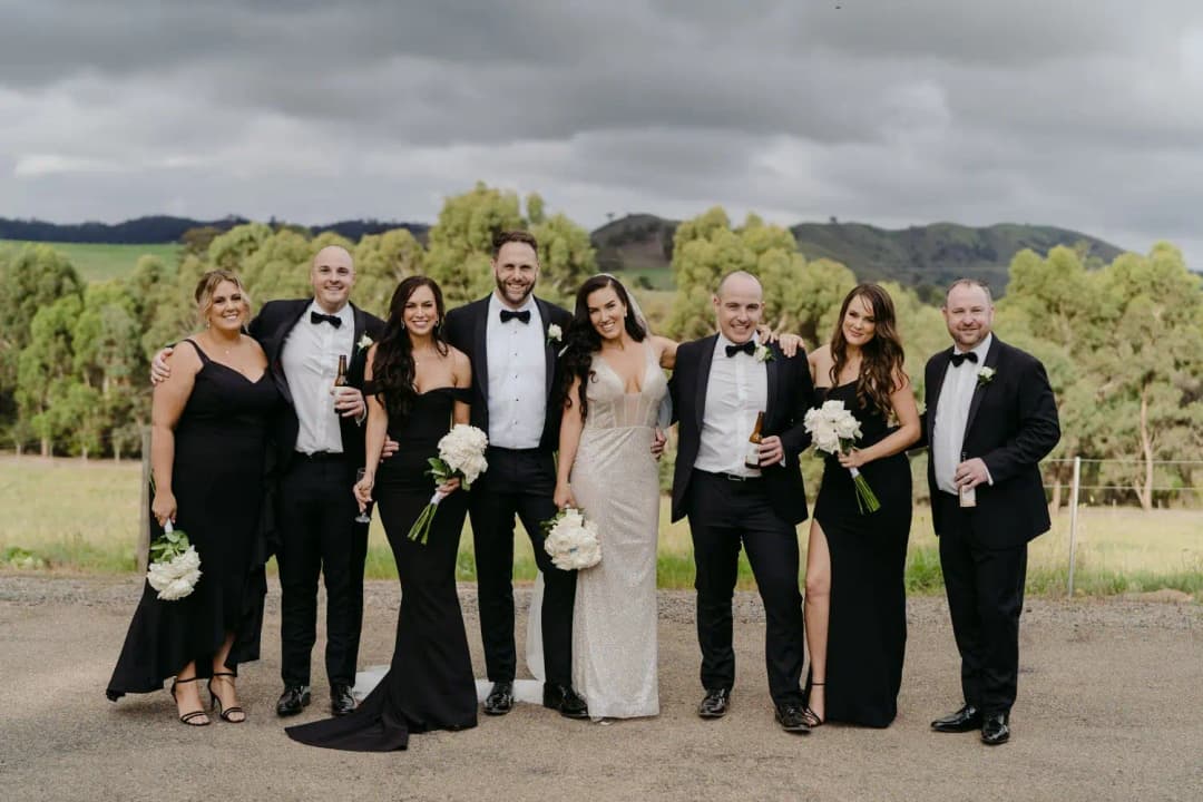 Couple poses with wedding guests against a vineyard and cloudy sky.