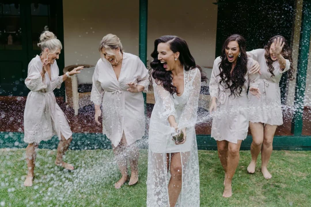 Women in robes laugh as champagne sprays across a lawn.
