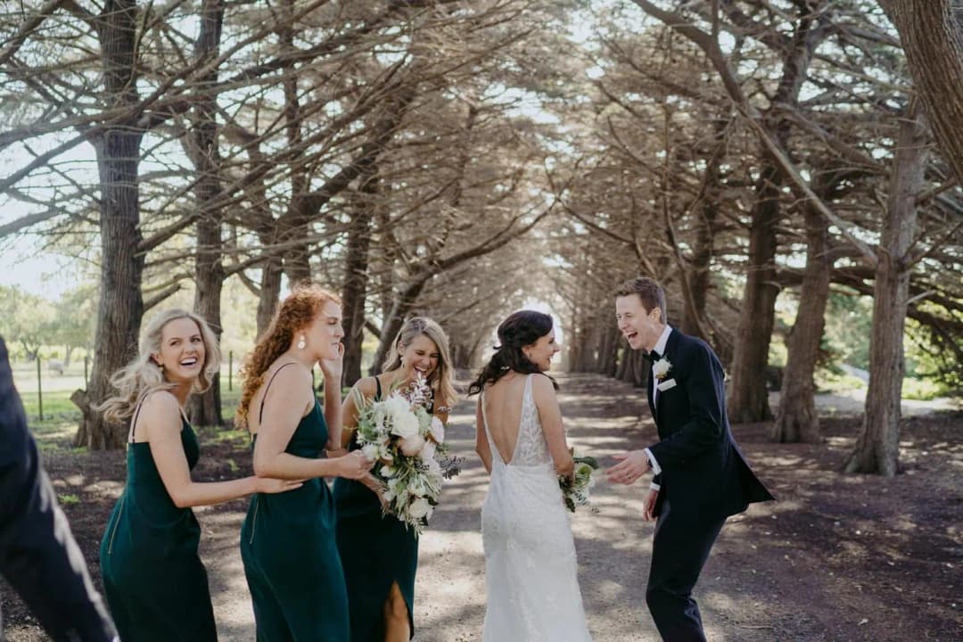 Couple and bridesmaids laugh together beneath tall trees.