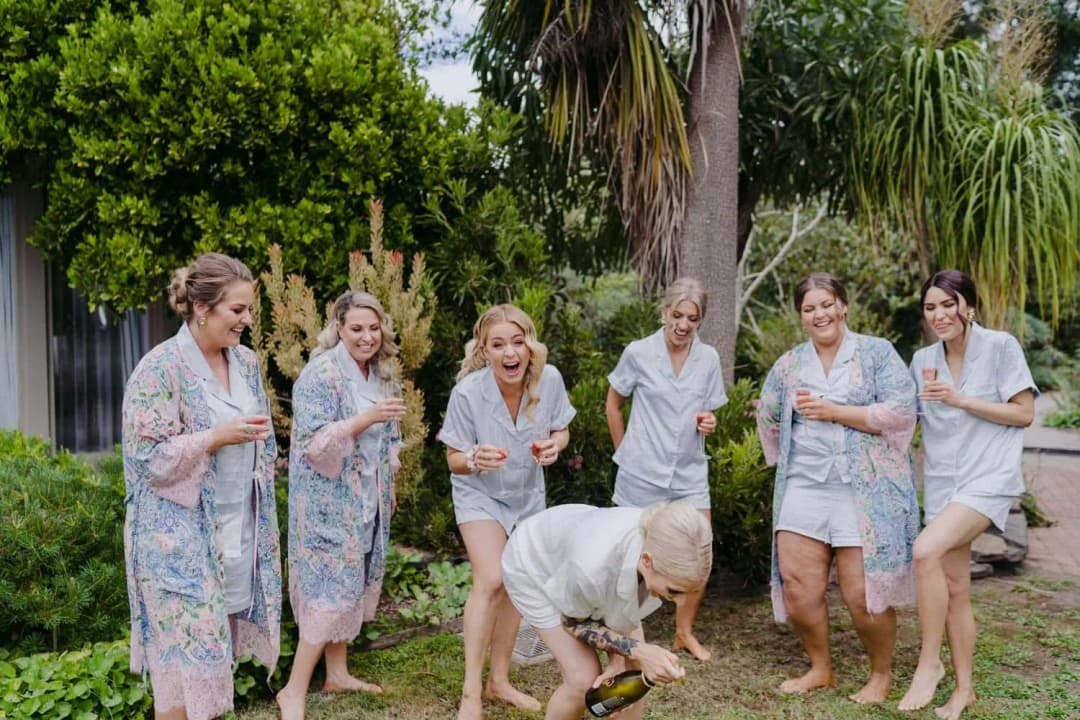 Women in robes and pyjamas laugh as a champagne bottle is opened close to the ground.