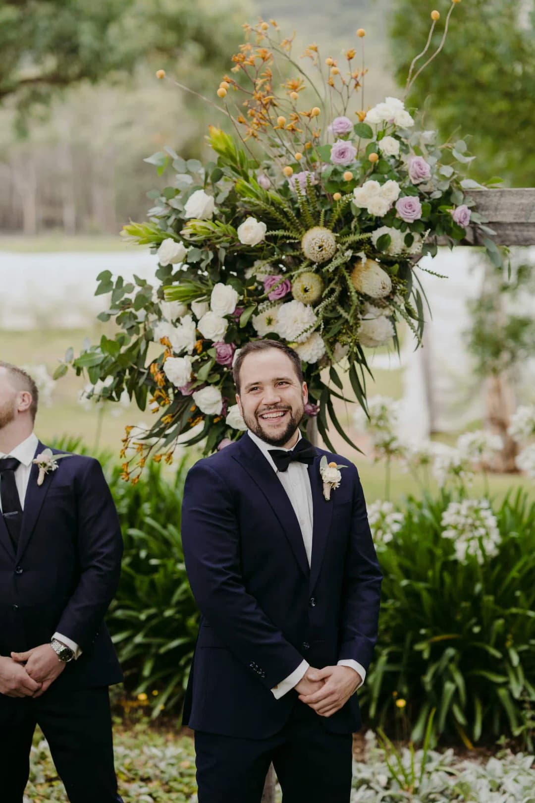 Groom smiles while waiting beside a tall floral arrangement.