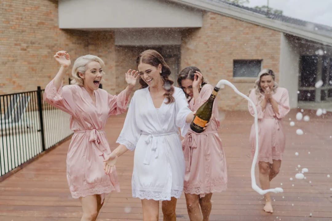 Women in pink and white robes laugh as champagne sprays on a terrace.