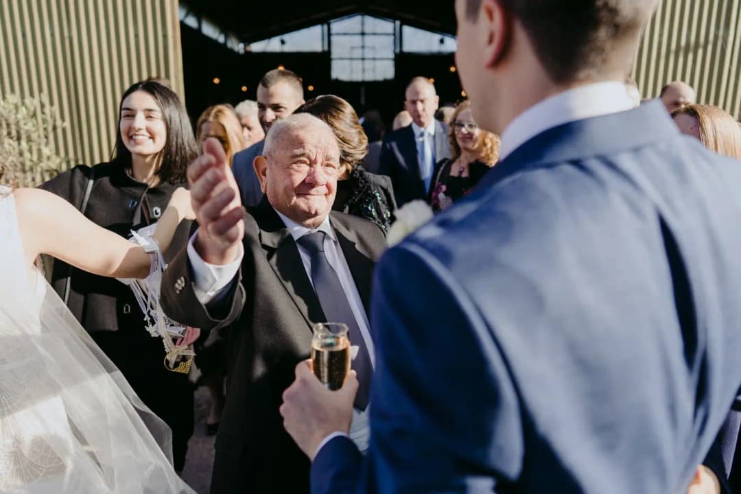 Older guest reaches toward the couple while holding a champagne glass.
