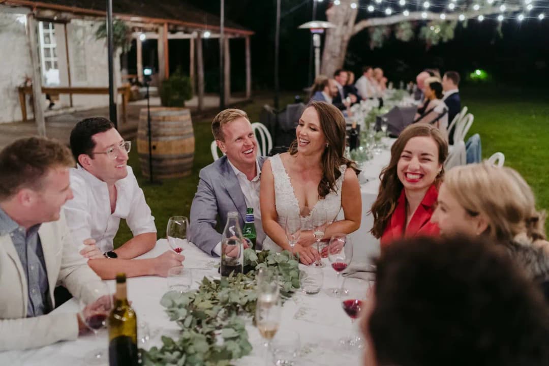 Bride and guests laugh together at an outdoor reception table beneath string lights.