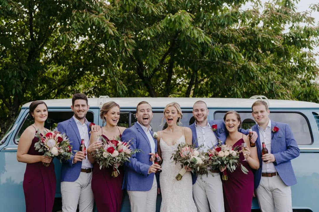 Wedding party smiles beside a blue van while holding bouquets and drinks.