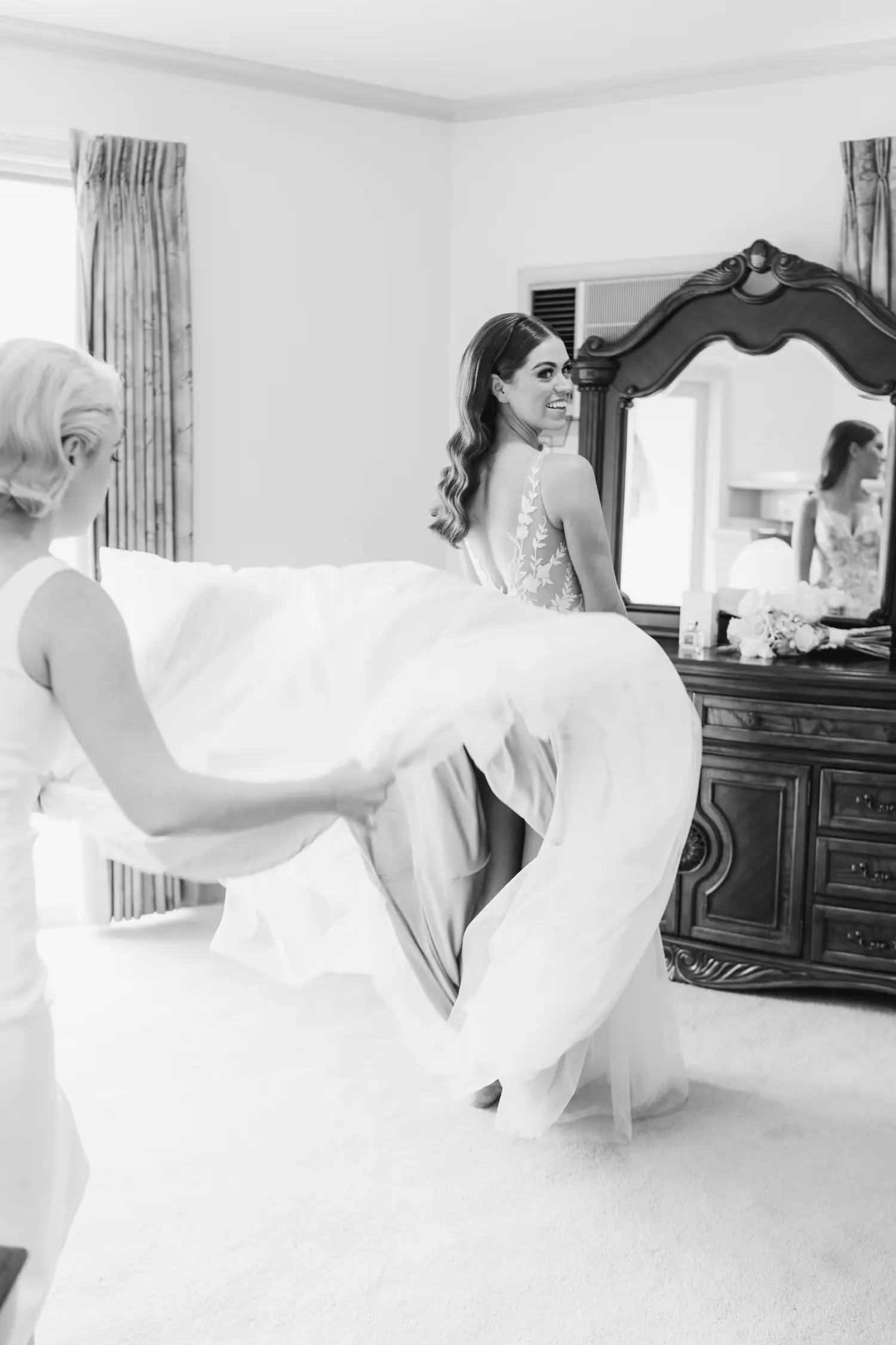 Black and white photograph of a friend holding the bride's dress train indoors.