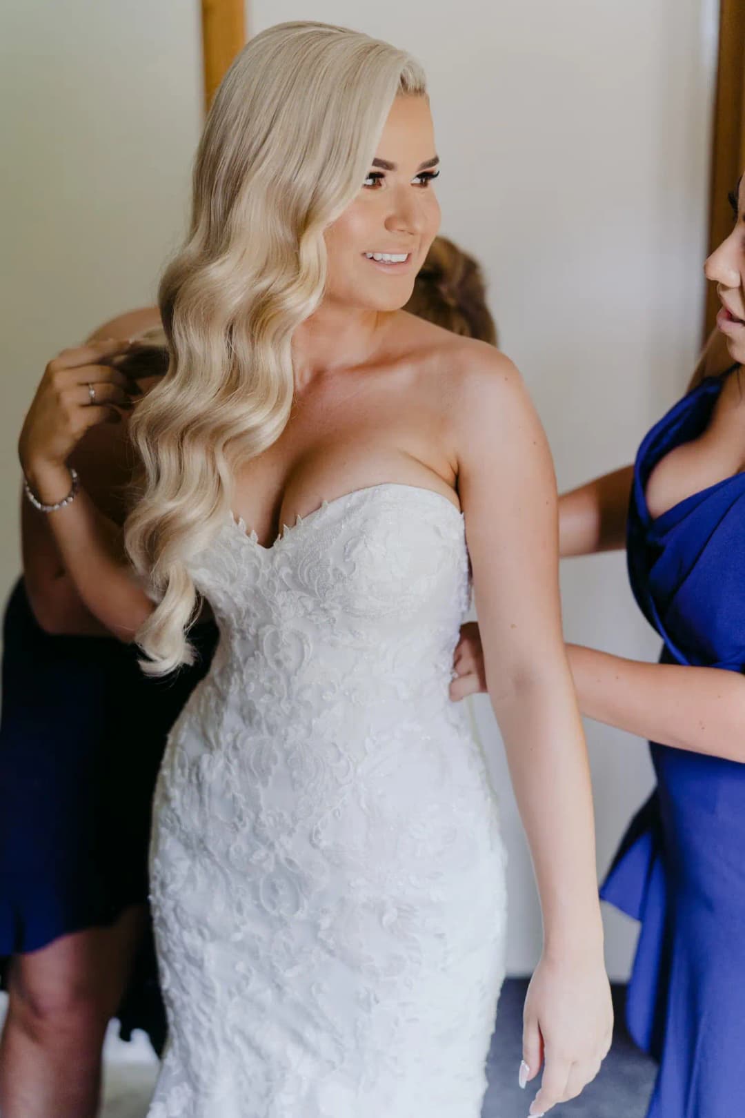 Bride smiles toward a friend as her dress is adjusted.