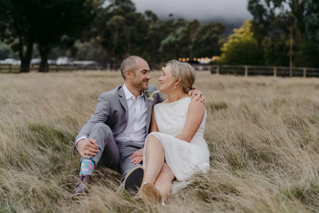 Couple sits together in long grass with their arms around each other.