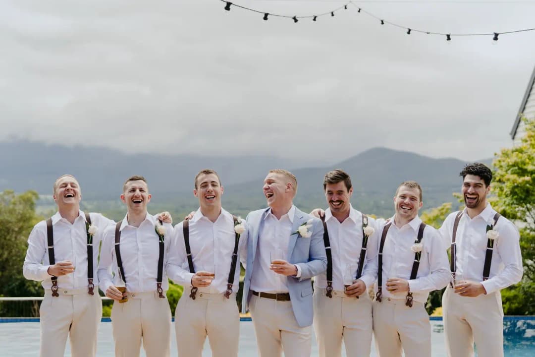 Men in white shirts and braces laugh together while holding drinks outdoors.