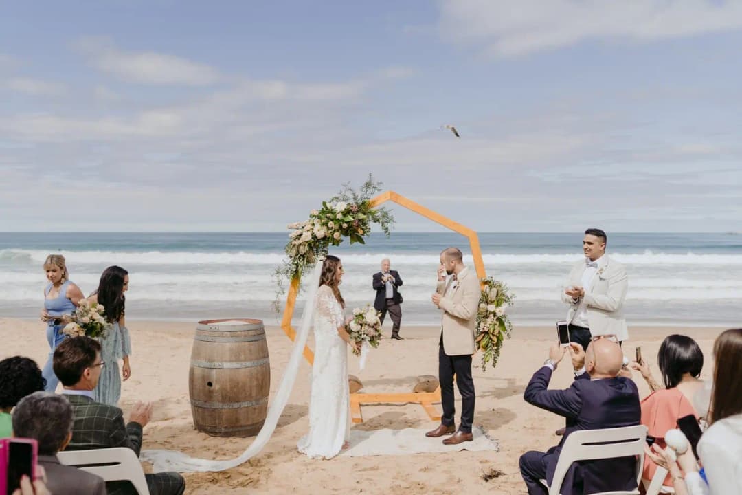 Beach ceremony beneath a decorated wooden arch with the ocean behind the couple.