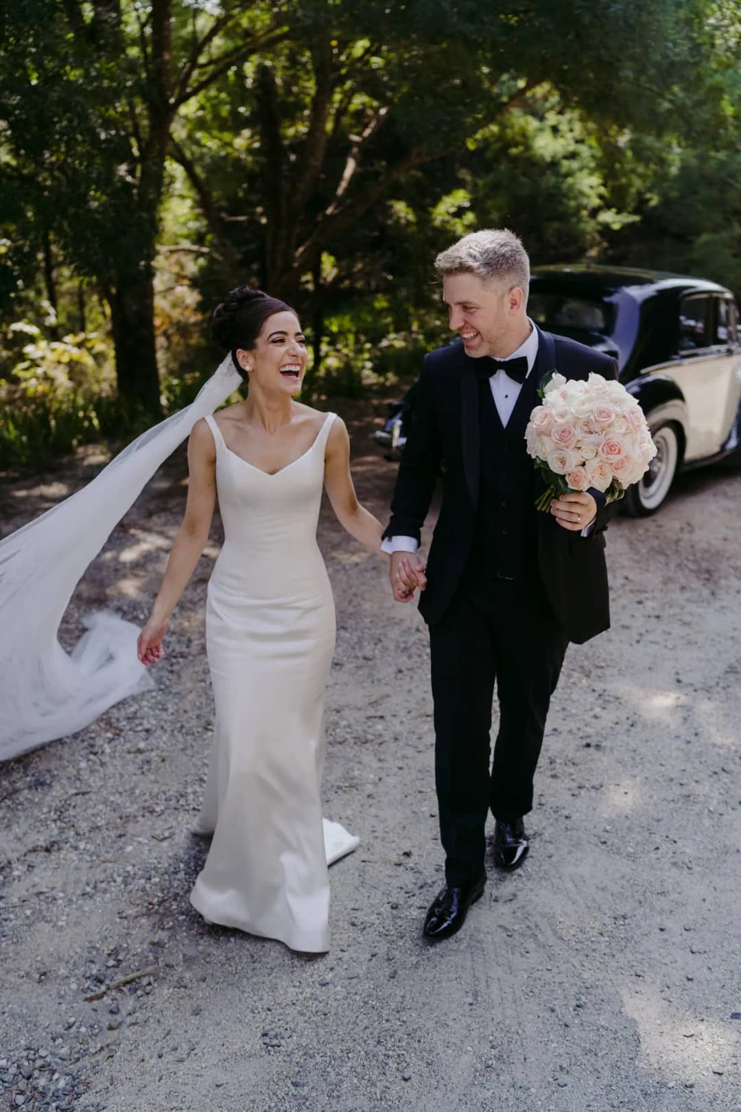 Couple walks along a tree-lined gravel road holding a bouquet.