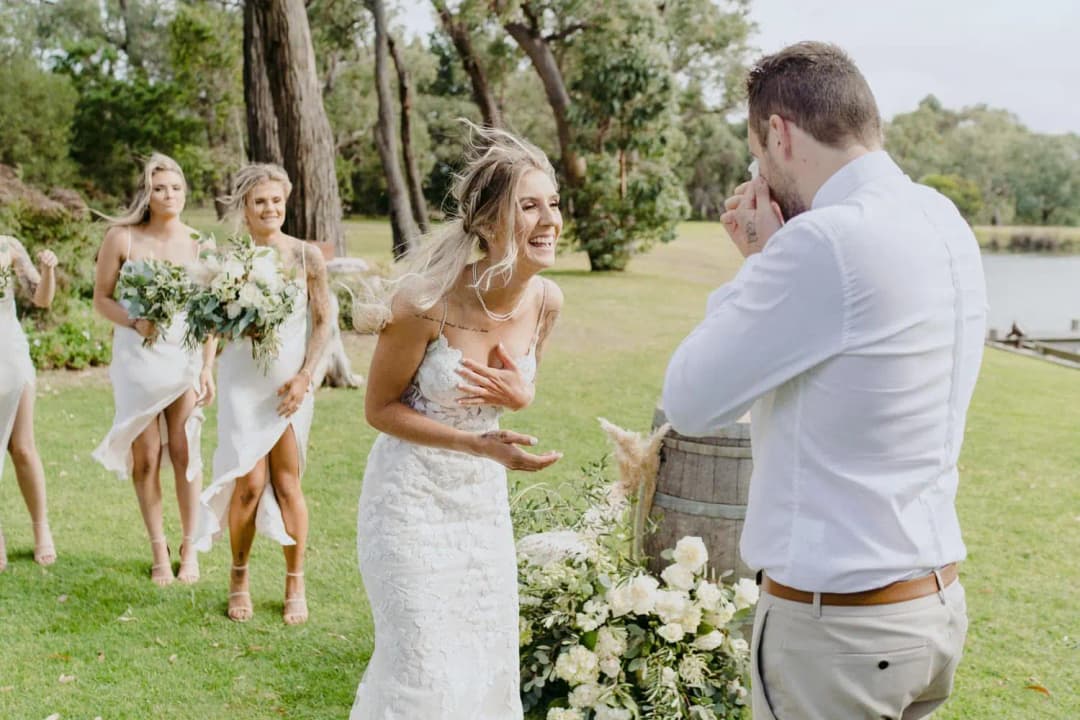 Bride laughs as the groom wipes his eye beside a pond.